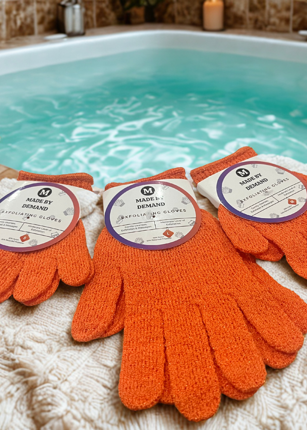 Orange exfoliating gloves with 'Made by Demand' labels on a textured surface with a bathtub in the background.